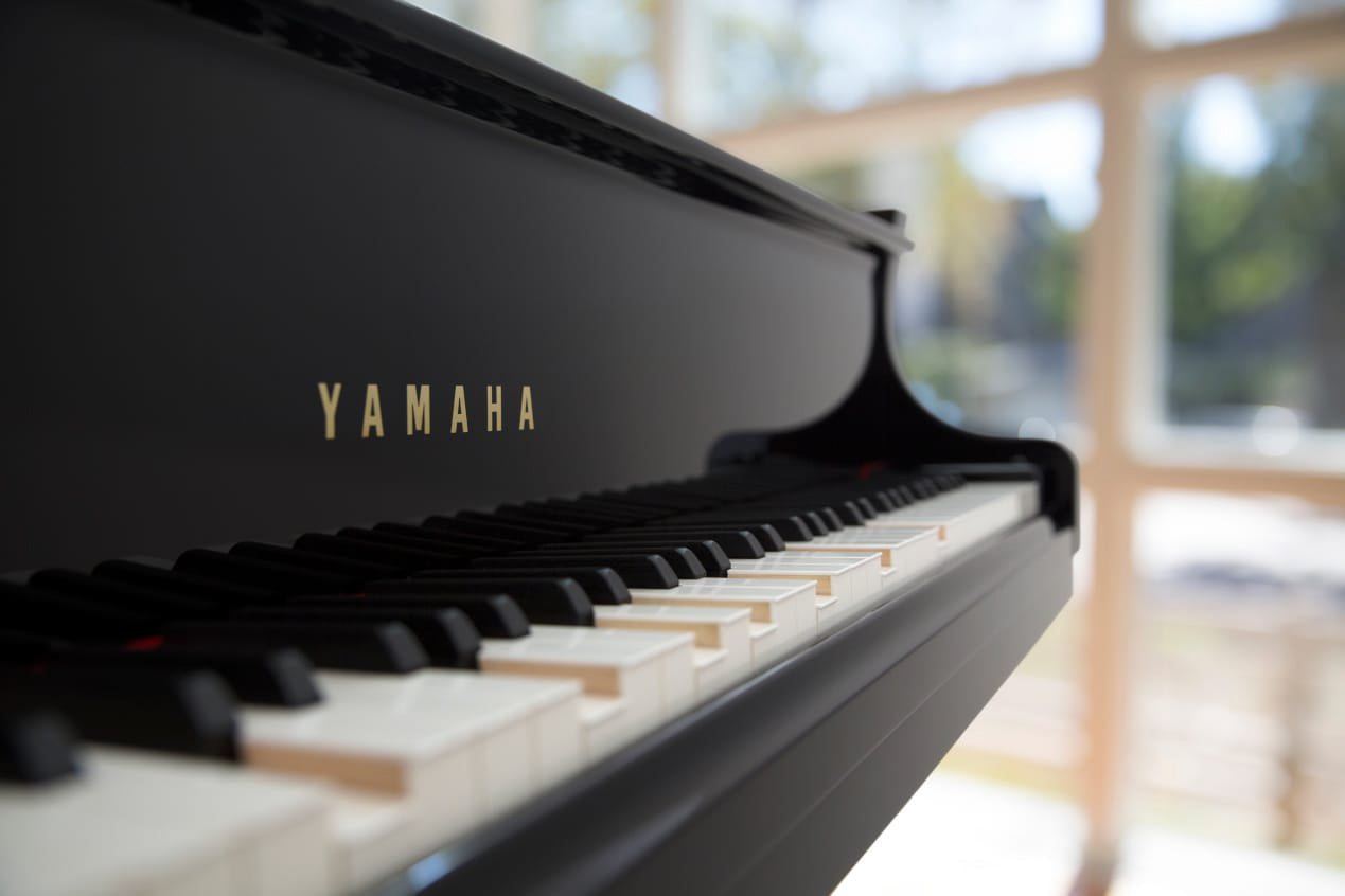 Close-up of a black Yamaha grand piano keyboard with focus on the brand logo, situated in a room with bright natural light coming through windows in the background.