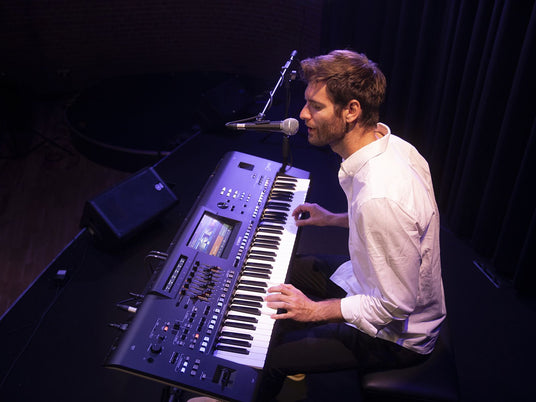 A male musician playing a digital keyboard on stage with a microphone in front of him, illustrating a modern piano performance.