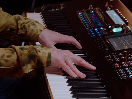 A musician's hands playing on a modern electronic keyboard with various control knobs and an LCD screen.