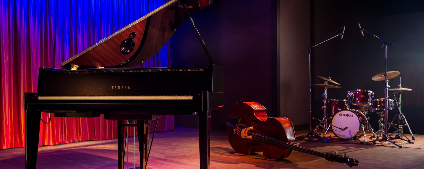 Grand piano on a stage with red curtain background along with a cello and drum set, representing a classical and contemporary fusion music setup.