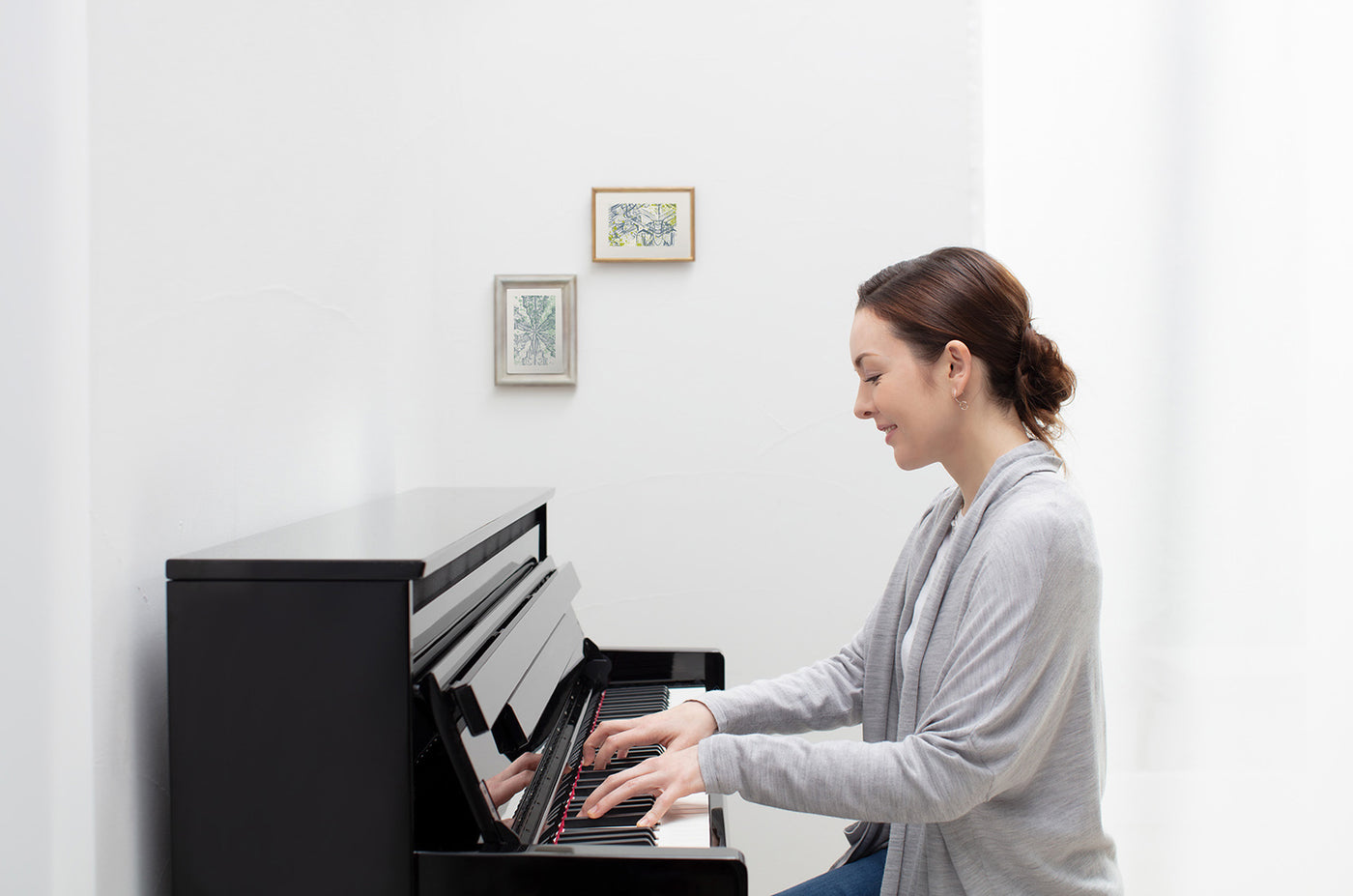 Woman practicing on a modern upright piano in a brightly lit room with framed artwork on the wall.