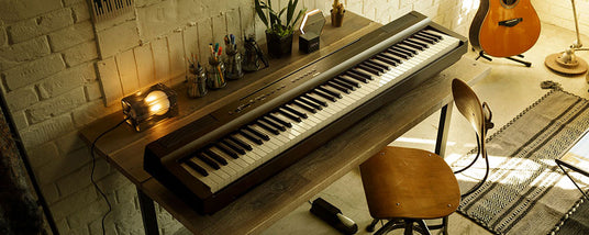 Cozy home music studio setup with an electronic piano on a wooden desk, illuminated by a warm desk lamp, with a guitar leaning against the wall, an artistic chair, and decorative items on shelves against a brick wall background.