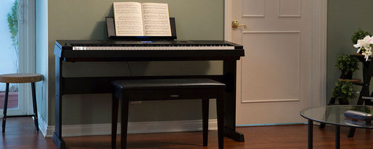A digital piano with sheet music on the stand, placed in a cozy room with hardwood floors, next to a bench, with a side stool and a glass coffee table in the foreground.