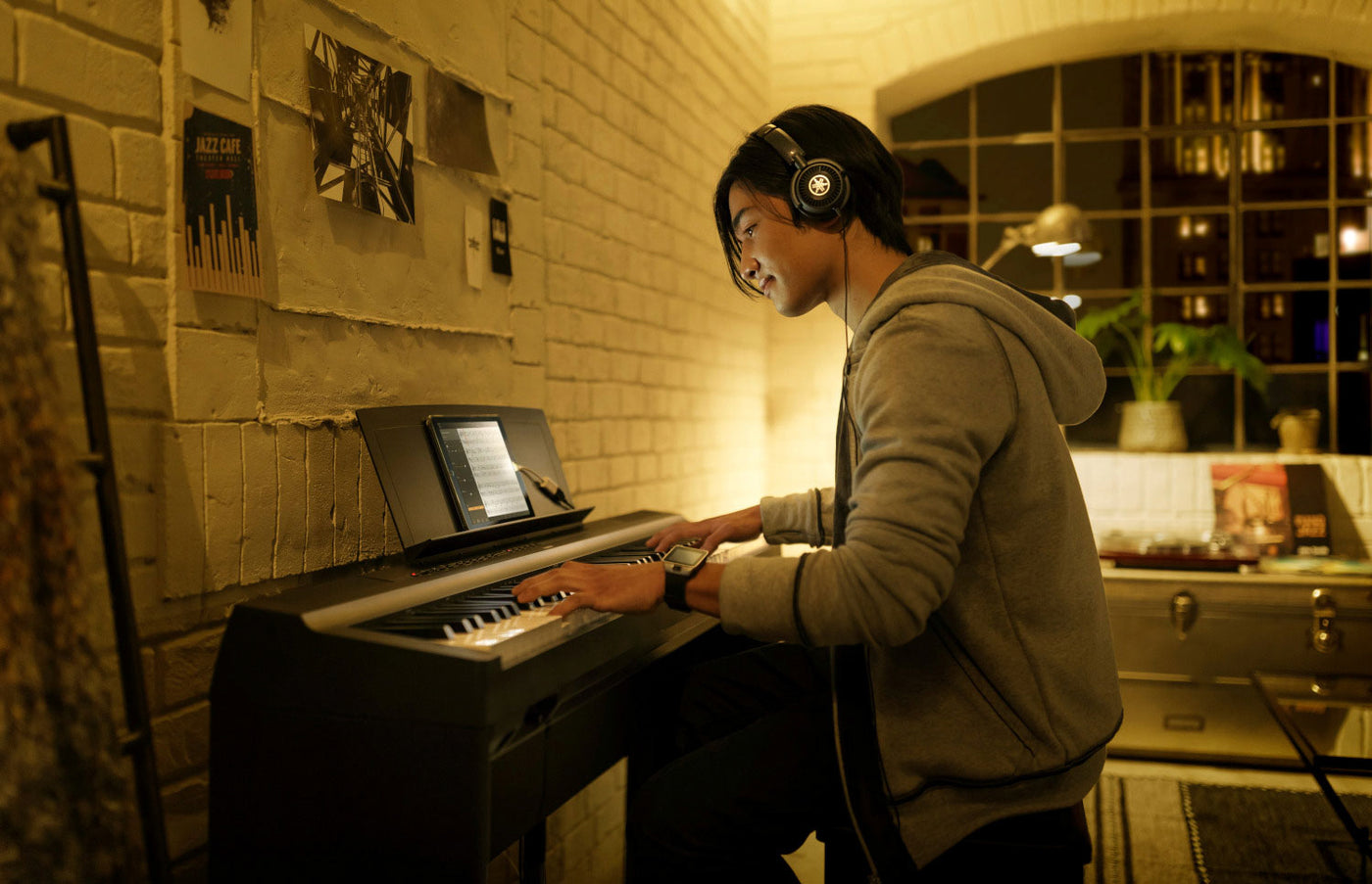 A person wearing headphones playing an electronic piano with a digital sheet music display in a cozy room with brick walls and warm lighting.