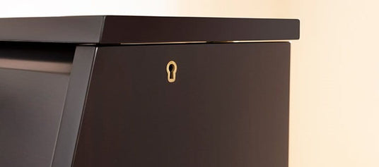 Close-up of a piano's top board, focusing on the lock mechanism. The image shows the edge of the dark, polished top board and part of the piano's upper panel. A small, gold-colored keyhole for the top board lock is centered in the frame.