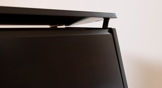 Close-up view of a piano's top board slightly raised, showing the tone escape feature. The image captures the edge of the black piano cabinet with the top lid lifted a small amount.