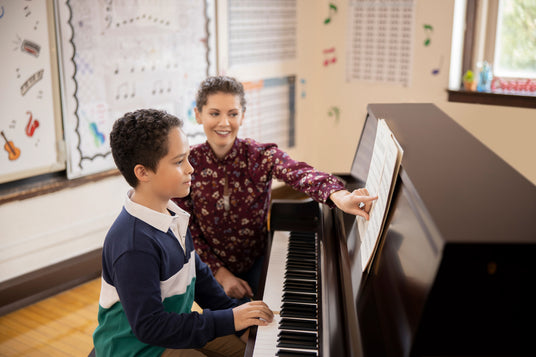 Young student sitting at a piano and being guided by a teacher who is pointing to sheet music during a piano lesson.