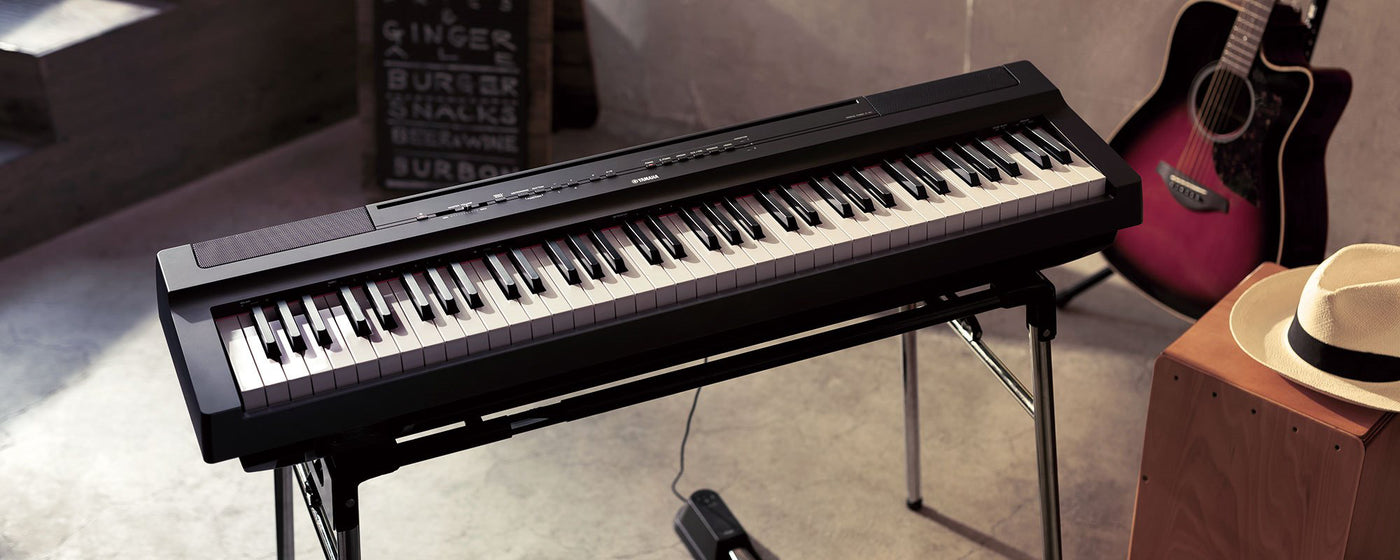 Black digital stage piano on a stand showcased in an urban rehearsal space with a red acoustic guitar and a straw hat on wooden furniture, reflecting a casual music-making setting.