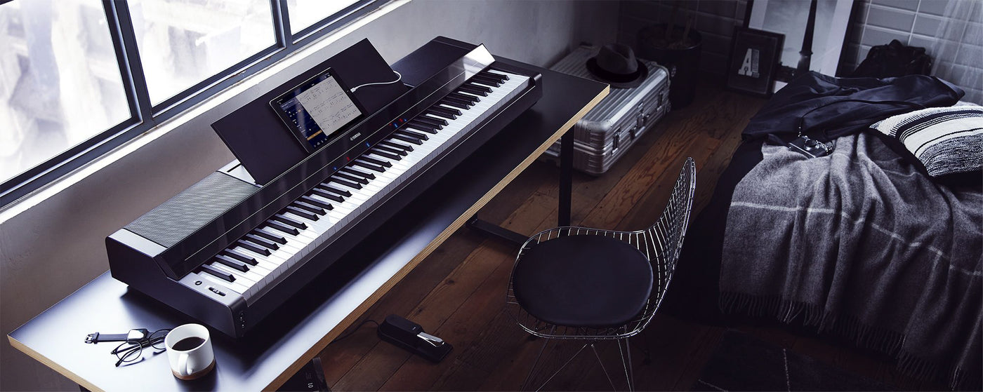 Modern digital piano with weighted keys by a window in a cozy interior setup, equipped with a tablet displaying sheet music, headphones, and a sustain pedal.