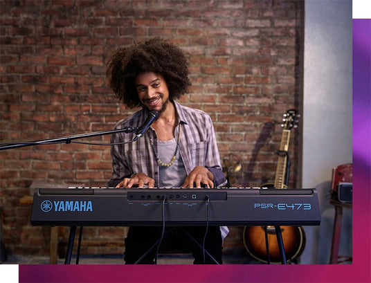 A smiling musician playing a Yamaha PSR-E473 keyboard in a room with a brick wall background, microphone stand in front, and an acoustic guitar in the background.