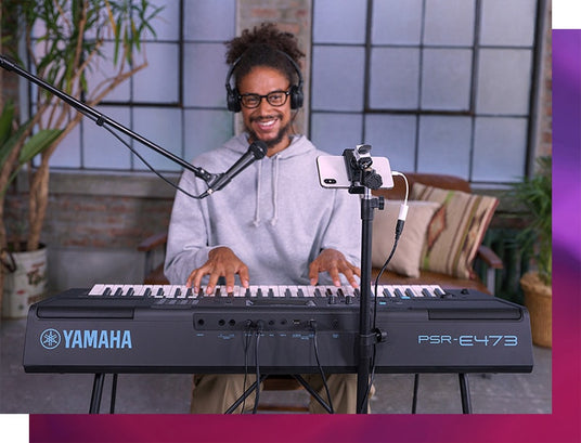 A smiling musician wearing headphones playing a Yamaha PSR-E473 keyboard with a microphone and smartphone attached to a tripod in a cozy room with brick walls and plants.