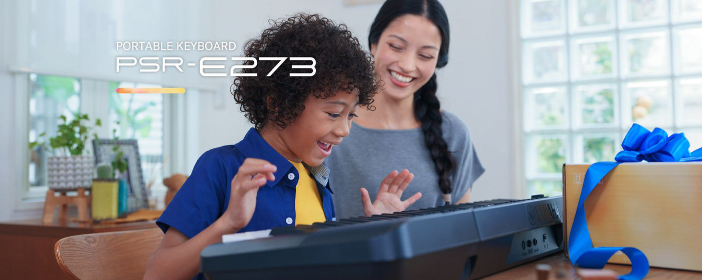 Young child happily playing on a PSR-E273 portable keyboard while a smiling adult woman watches, with a gift-wrapped box nearby, suggesting a music-related present in a home setting.