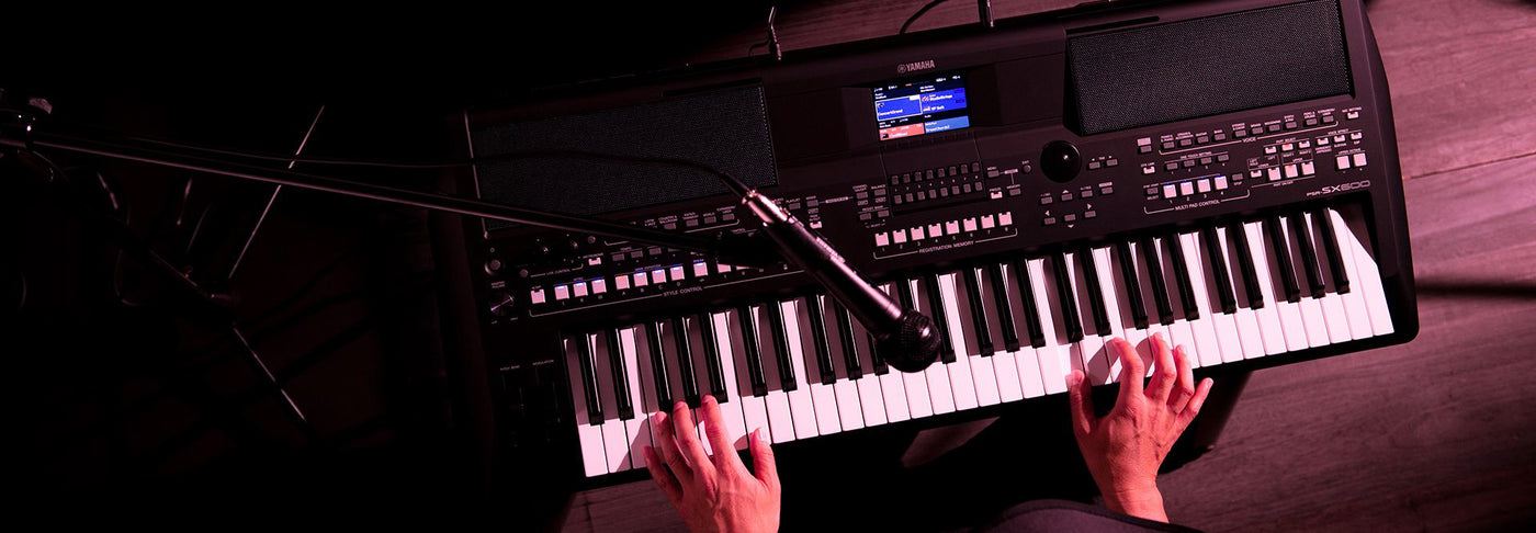 A close-up view of hands playing a Yamaha PSR-SX900 digital keyboard with an attached microphone, illustrating a modern musical performance setup.