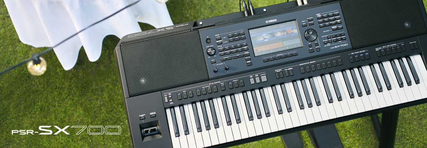 Close-up view of a Yamaha PSR-SX700 electronic keyboard with focus on the keys, buttons, and digital display, set up outdoors near a white draped table with a hanging light bulb.