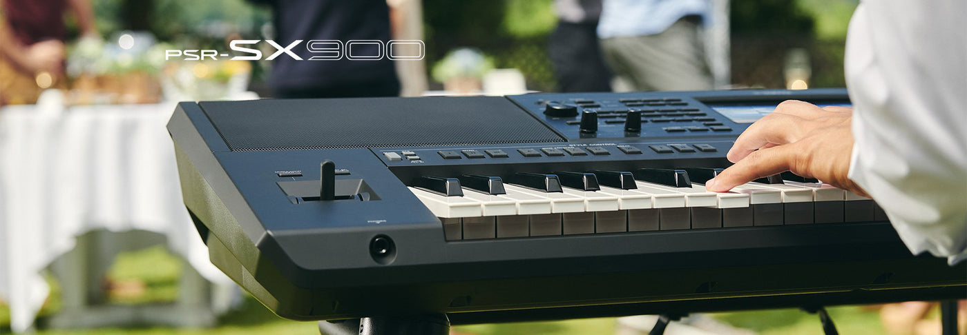 A person playing a black PSR-SX900 digital keyboard at an outdoor event with a blurred background of guests and tables.