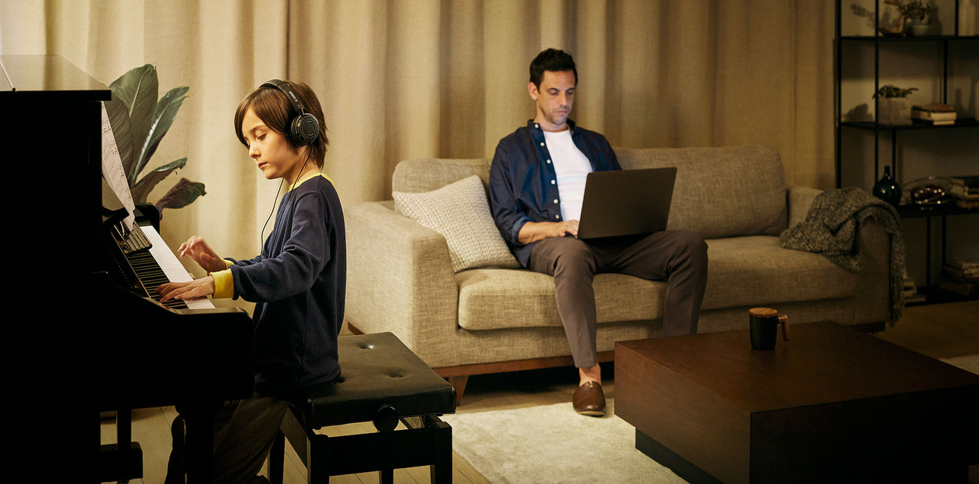 A young person practicing on a digital piano with headphones in a cozy living room while another individual works on a laptop on the couch, illustrating a quiet environment for music practice with a modern piano that caters to both learning and respectful cohabitation.