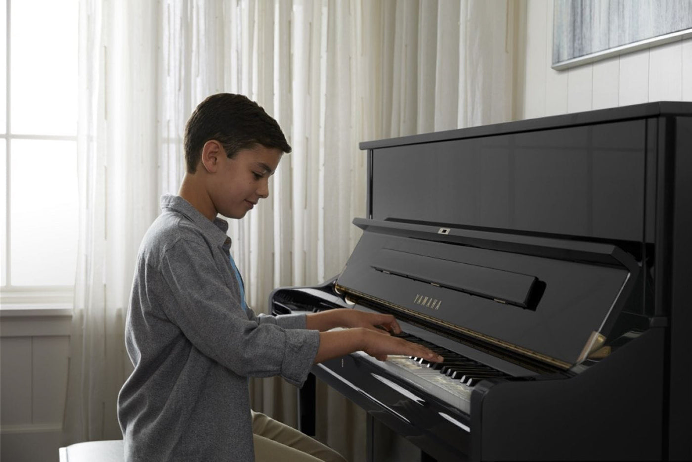 A young person practicing on a black Yamaha upright piano in a room with natural light filtering through sheer curtains.