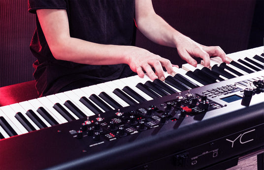 A musician playing a modern digital stage piano with various controls and sliders visible, set against a red-tinged backdrop suggestive of a live performance or recording session.
