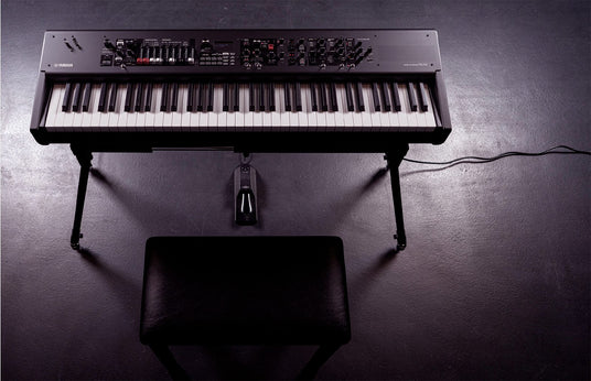 A Yamaha synthesizer with a full set of keys placed on a stand over a dark surface, accompanied by a sustain pedal and a stool, with cables trailing on the floor.