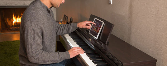 Man in a grey sweater playing a digital piano with a tablet on the music stand in a cozy room with a fireplace in the background.