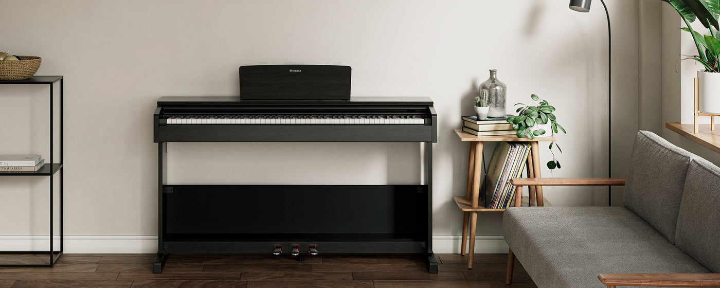 A modern black digital piano placed against a neutral wall in a minimalist living room with decorative plants and books, showcasing the integration of musical instruments with home decor.
