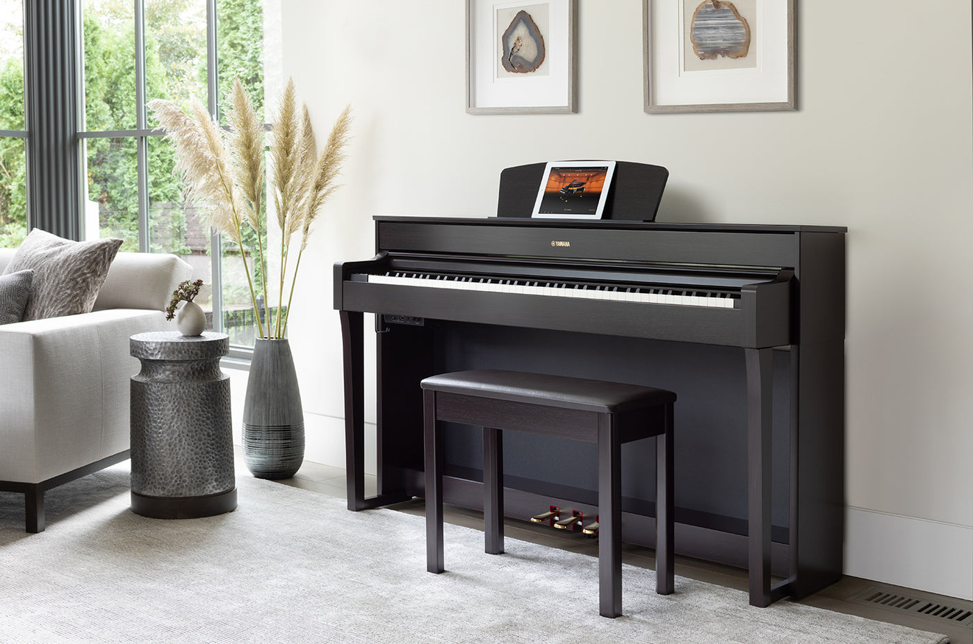 A modern black upright piano with a matching piano stool set in a bright room with large windows, framed wall art, and decorative elements, exemplifying a contemporary home setting.