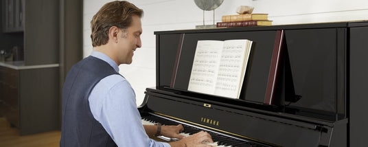 Man seated at a black Yamaha upright piano, playing from a sheet music book in a home setting.