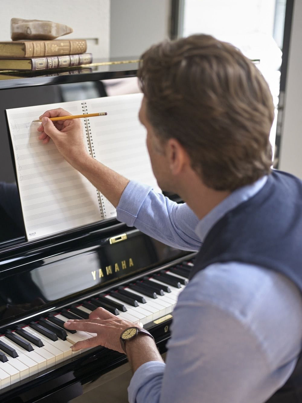 Composer sitting at a Yamaha piano, writing music on a staff paper with a pencil.